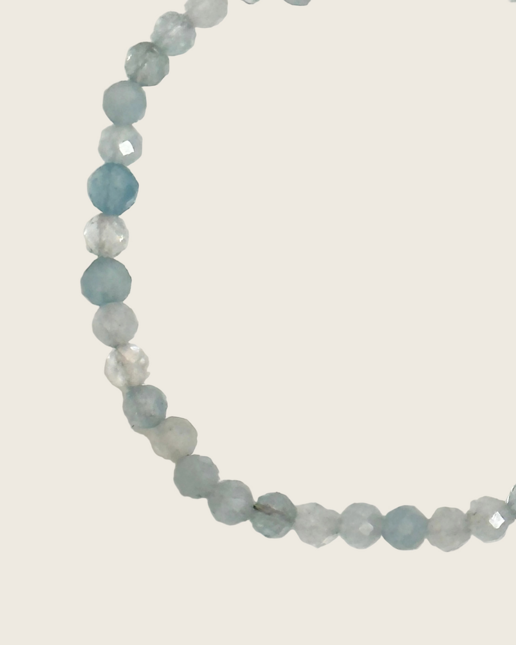 A beaded strand of faceted light blue and white gemstone beads, arranged in a curved shape on a plain off-white background.