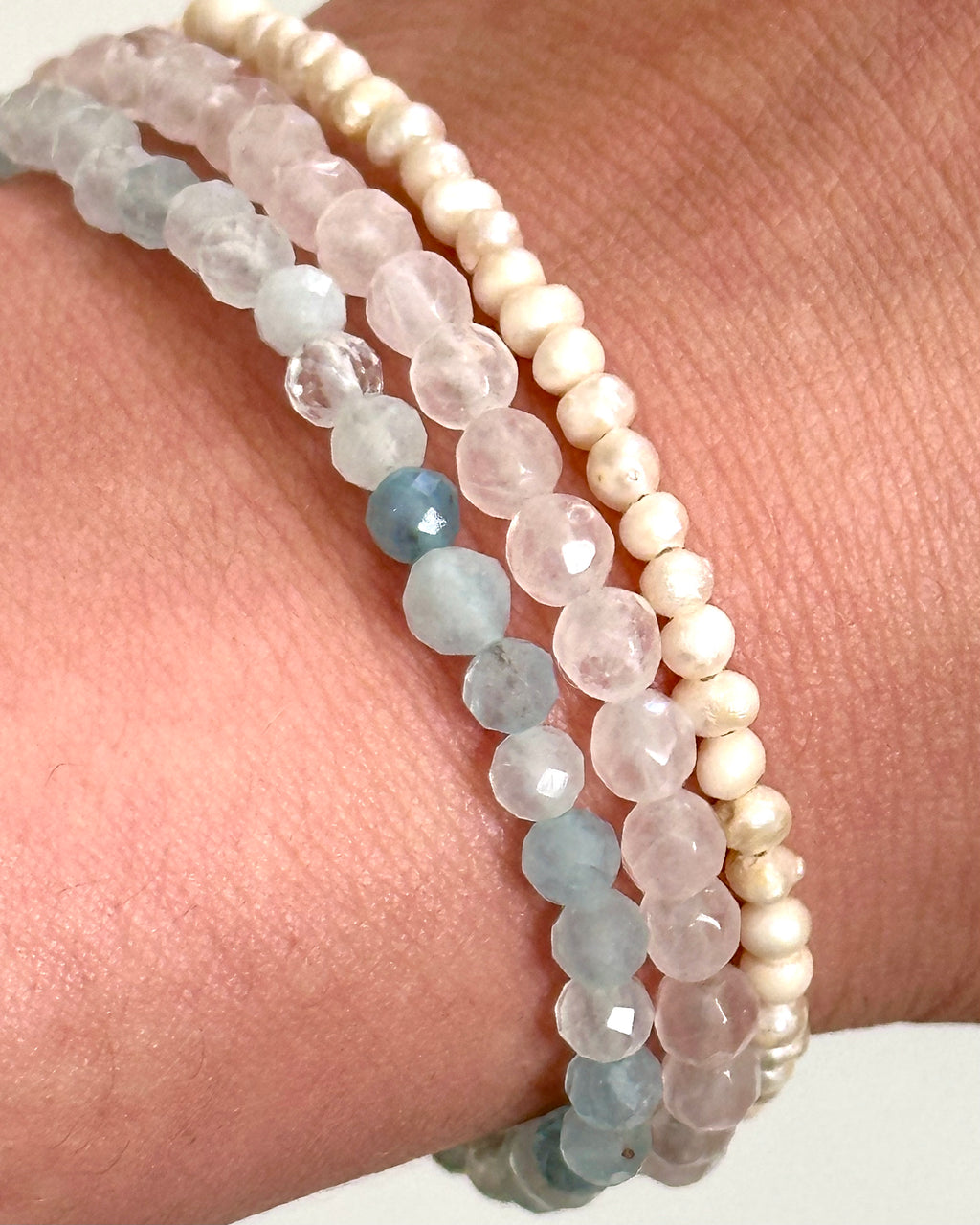 Three beaded stretch bracelets on a wrist: one strand of cream beads, one of pale pink faceted beads, and one of light blue faceted beads.