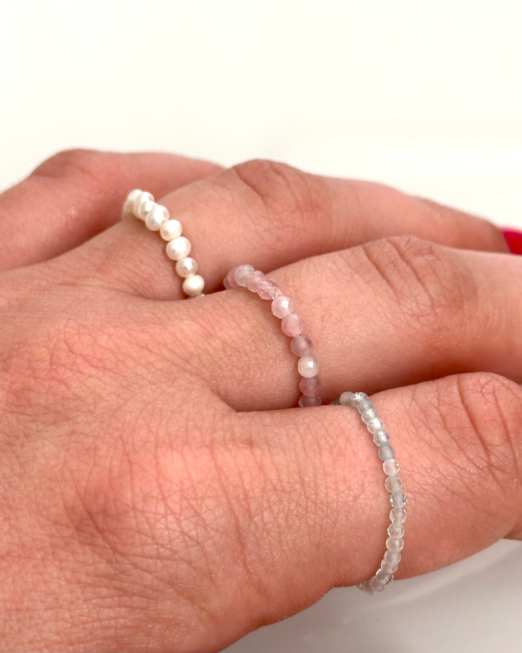 Three delicate beaded rings displayed on a person's hand: a white pearl bead ring, a pale pink stone bead ring, and a light blue stone bead ring.