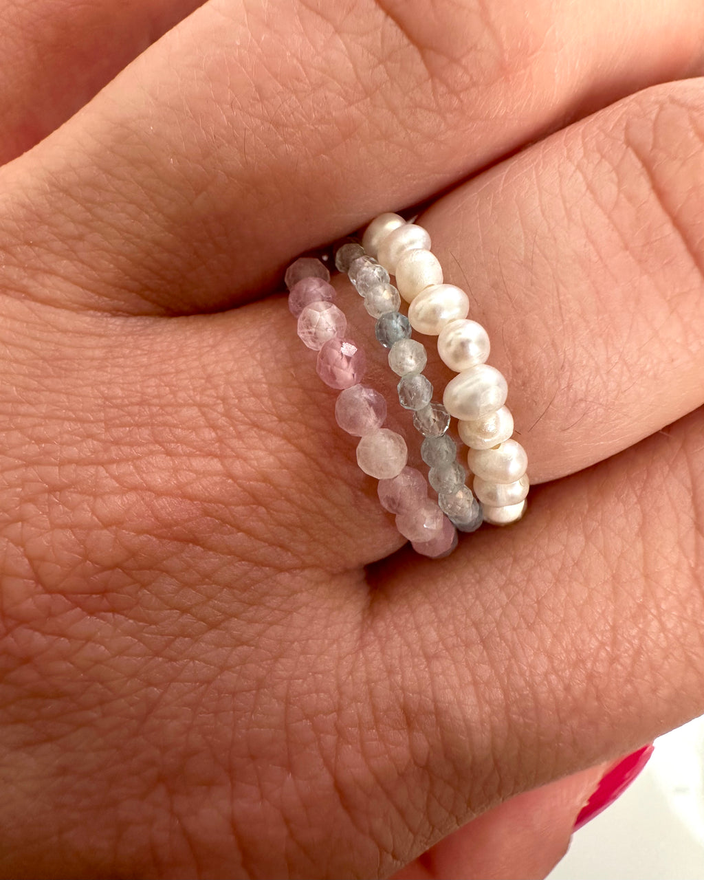 Three beaded stretch rings worn on a person's finger. The rings feature faceted pink beads, smooth white pearl-like beads, and translucent light gray beads.